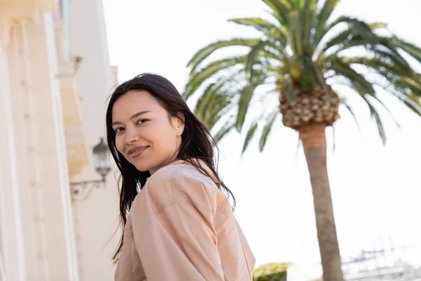 Happy woman looking at camera near blurred palm tree on street — стоковое фото