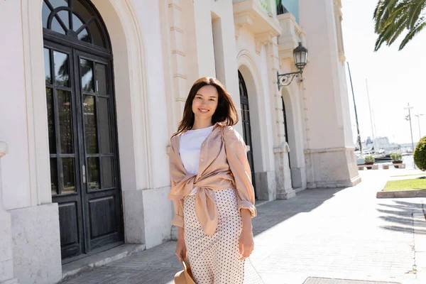 Mulher morena feliz e elegante olhando para a câmera perto do edifício branco com portas pretas — Fotografia de Stock