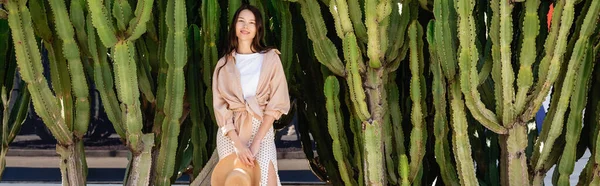 Cheerful woman in stylish clothes looking at camera near green giant cacti, banner — Stock Photo