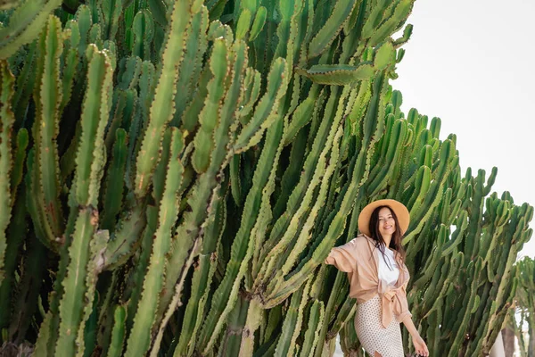 Fröhliche Frau in stylischer Kleidung und Strohhut im Park mit riesigen Kakteen — Stockfoto