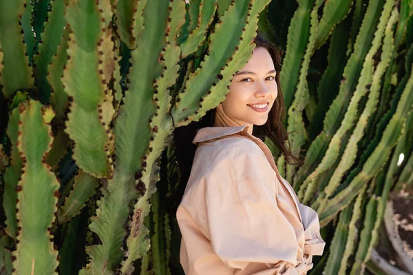 Happy woman in beige shirt looking at camera near green cacti — Stock Photo