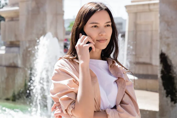 Brünette Frau schaut weg, während sie am Handy in der Nähe des verschwommenen Brunnens spricht — Stockfoto