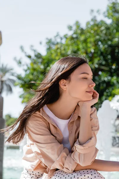 Dreamy and positive brunette woman holding hand near face while sitting outdoors — Foto stock