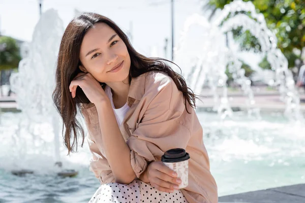 Mulher sorrindo em camisa bege segurando café para ir e sorrindo para a câmera perto de fonte turva — Fotografia de Stock