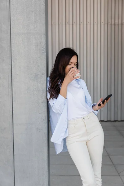 Brunette woman drinking coffee and using smartphone near grey wall — Stockfoto