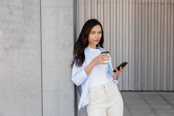 Brunette woman with takeaway drink and mobile phone looking at camera near grey wall — Stockfoto