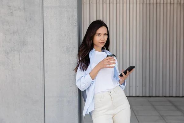 Mulher morena com bebida takeaway e telefone celular olhando para a câmera perto da parede cinza — Fotografia de Stock