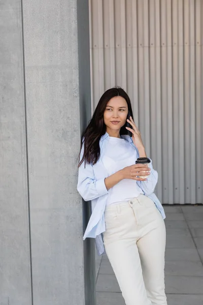 Женщина в белых брюках держит кофе, чтобы пойти во время разговора на смартфоне на открытом воздухе — стоковое фото