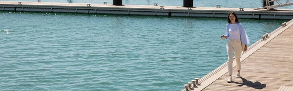 Full length of brunette woman with paper cup walking along pier, banner — Stock Photo