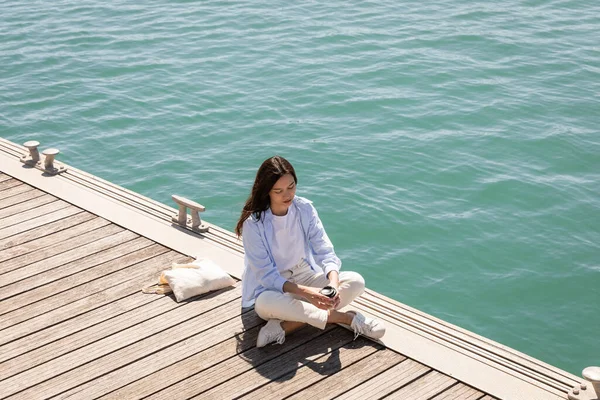 High angle view of woman holding paper cup while sitting on pier near canvas bag — Stock Photo