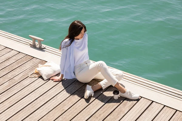 Hochwinkelaufnahme einer Frau, die auf einem Steg in der Nähe von Pappbecher und Leinentasche sitzt — Stockfoto