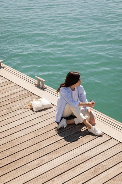 High angle view of brunette woman with coffee to go sitting on pier near canvas bag — Photo de stock
