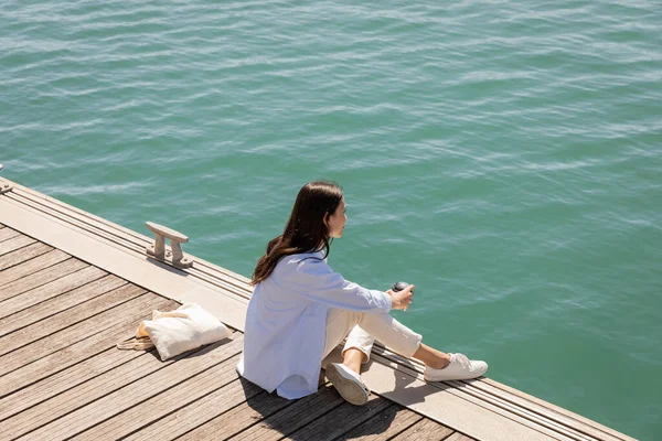 Vue grand angle de la femme assise sur la jetée avec du café pour aller et regarder la mer — Photo de stock