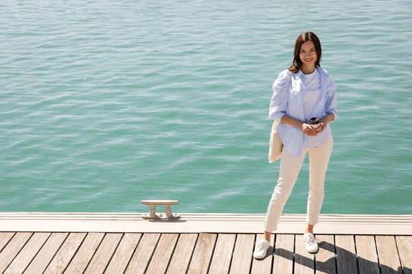 Full length of woman in blue shirt and white pants standing on pier with coffee to go - foto de stock