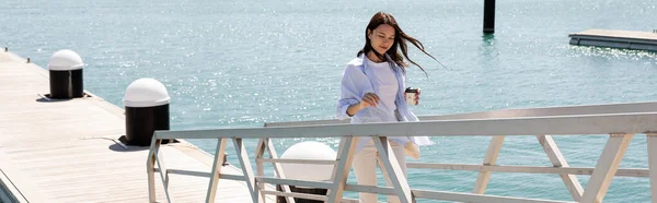 Femme brune avec boisson à emporter marchant sur la jetée de la mer, bannière — Photo de stock