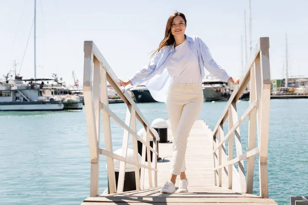 Volle Länge der lächelnden Frau in weißen Hosen, die auf der Seebrücke im Seehafen steht — Stockfoto