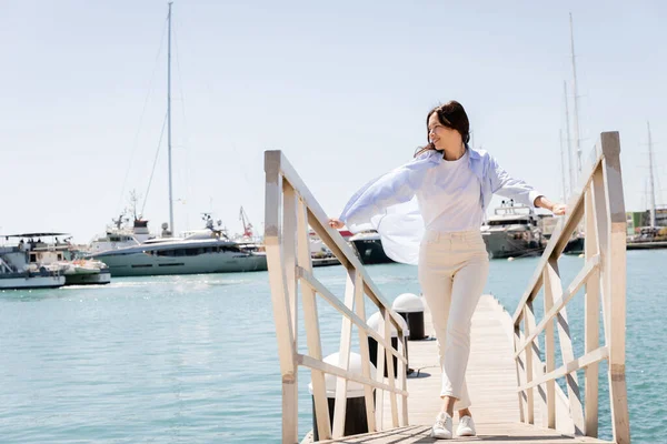 Full length view of joyful woman in white pants walking on pier in seaport — стоковое фото