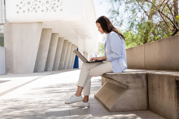 Side view of brunette woman in blue shirt and white pants using laptop outdoors — стоковое фото