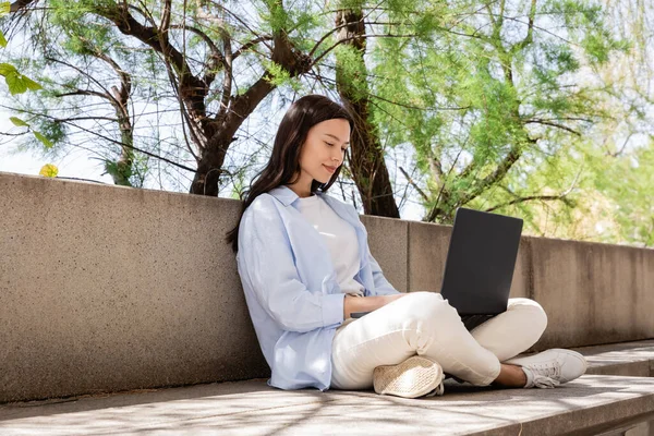 Full length of happy woman sitting with laptop on crossed legs outdoors — стоковое фото