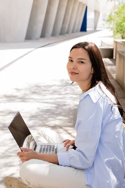 Happy brunette woman looking at camera near laptop with blank screen on urban street — Photo de stock