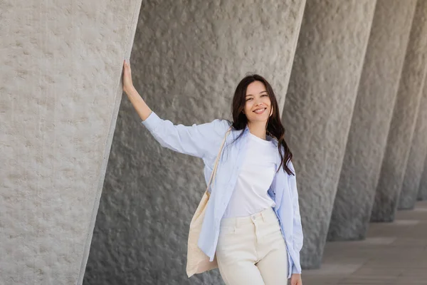 Happy brunette woman with canvas bag looking at camera near grey wall — Stock Photo