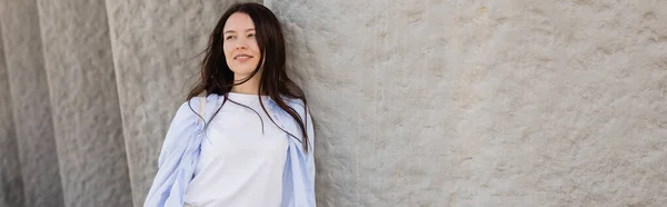 Femme brune en chemise bleue souriant et regardant loin près du mur gris, bannière — Photo de stock