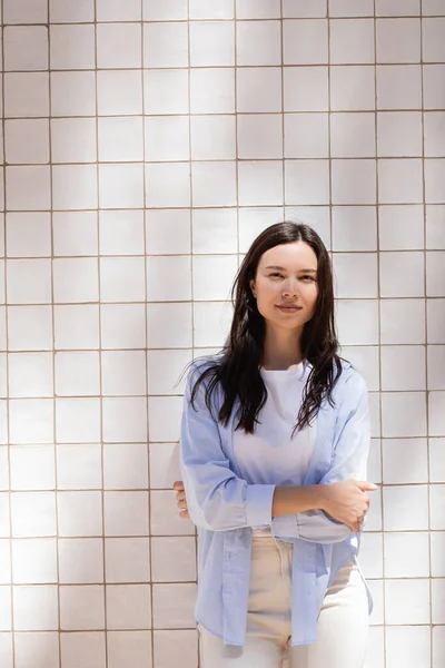Brunette woman in blue shirt standing with crossed arms near white tiled wall — Fotografia de Stock