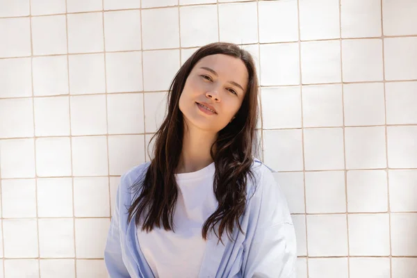 Femme brune souriante regardant la caméra tout en se tenant près du mur carrelé blanc — Photo de stock