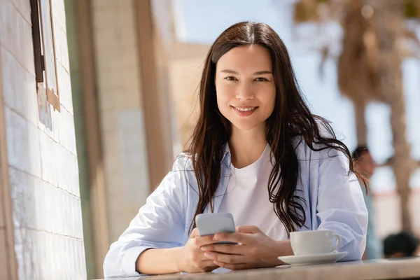 Joyful woman sitting with smartphone on summer terrace of cafe and looking at camera — Stock Photo