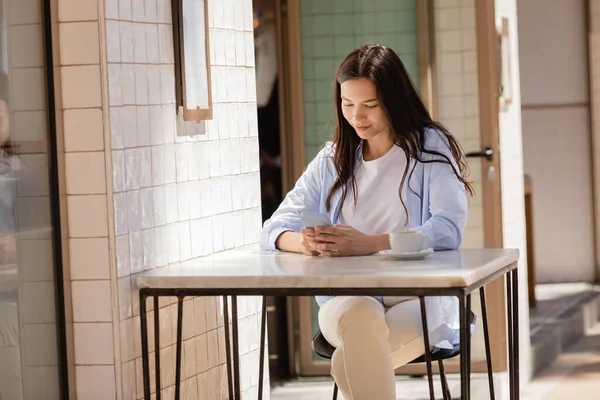 Smiling woman messaging on cellphone near coffee cup in street cafe — Foto stock