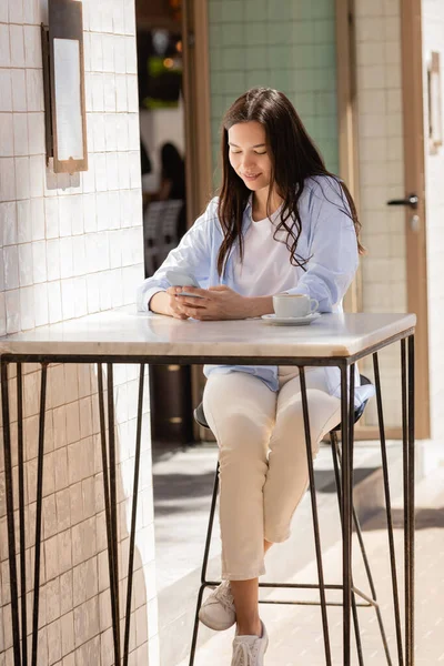 Happy brunette woman chatting on mobile phone near cup of coffee on cafe terrace — Stock Photo