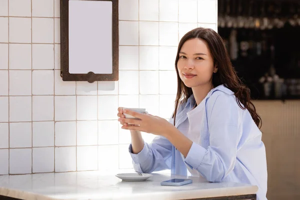 Brunette woman with cup of coffee looking at camera on cafe terrace — Stock Photo