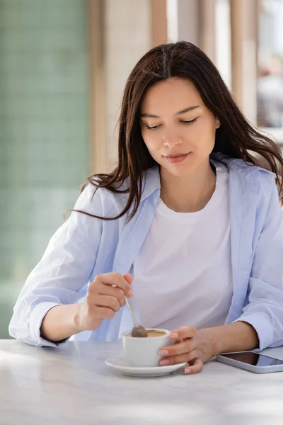 Brunette woman holding teaspoon near coffee cup and smartphone with blank screen on cafe terrace — Stockfoto