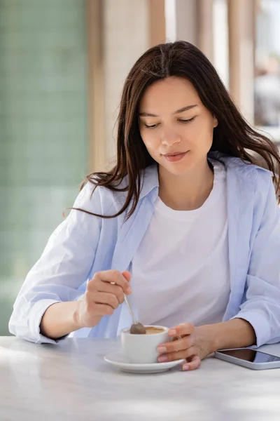 Brunette woman holding teaspoon near coffee cup and smartphone with blank screen on cafe terrace — Stock Photo