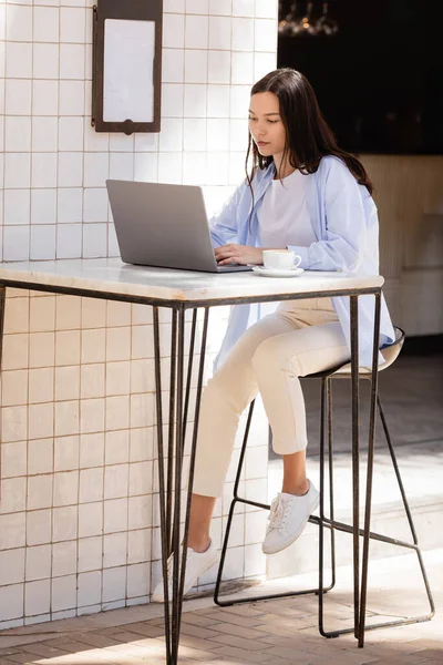 Vue pleine longueur de la femme brune tapant sur ordinateur portable près de tasse de café sur la terrasse du café — Photo de stock