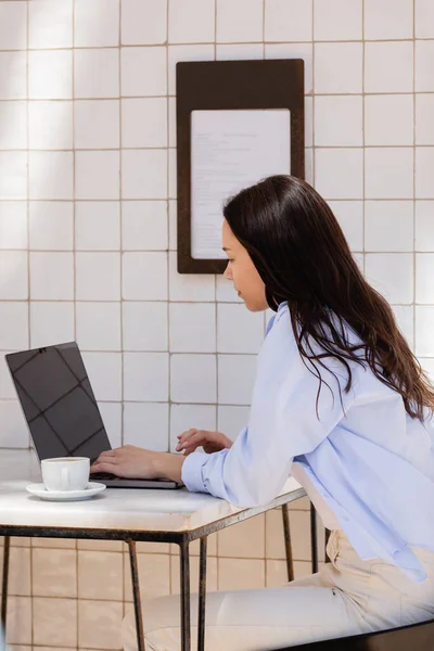 Вид сбоку на женщину рядом с ноутбуком с экраном и чашкой кофе на террасе кафе — стоковое фото