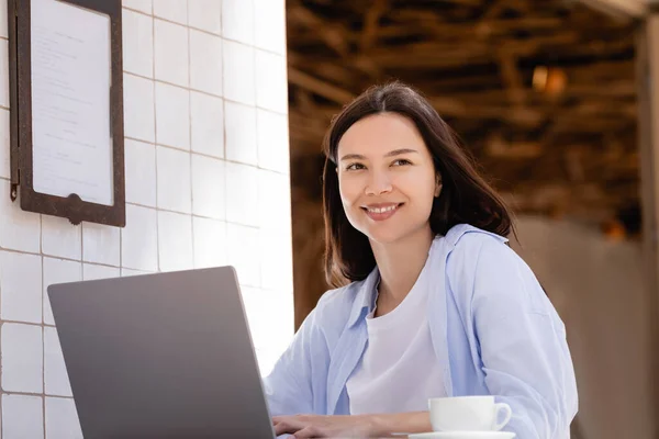 Cheerful woman smiling near laptop and coffee cup in street cafe on summer terrace — стоковое фото