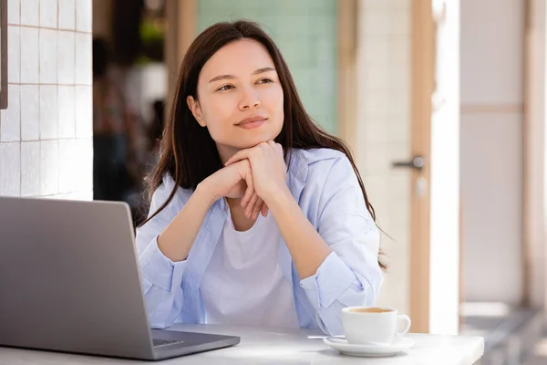 Мечтательная и позитивная женщина смотрит в сторону возле чашки кофе и ноутбука — стоковое фото