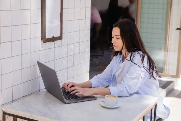 Woman in blue shirt typing on laptop near coffee cup in street cafe — Photo de stock