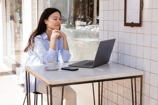 Улыбающаяся женщина смотрит на ноутбук рядом со смартфоном и чашкой кофе на летней террасе — стоковое фото