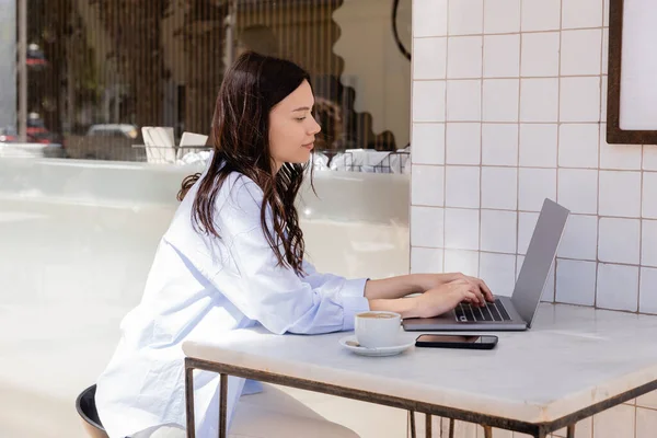 Вид сбоку женщины, сидящей на ноутбуке возле сотового телефона и чашки кофе в уличном кафе — стоковое фото