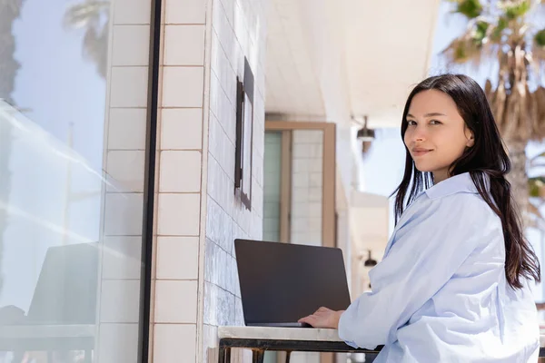 Mujer sonriente cerca de la computadora portátil con pantalla en blanco mirando a la cámara en la terraza del café - foto de stock