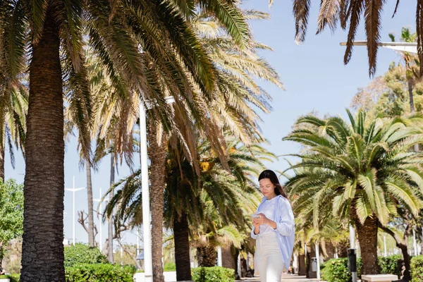 Femme brune debout sur une allée de palmiers dans le parc de la ville et utilisant un téléphone mobile — Photo de stock