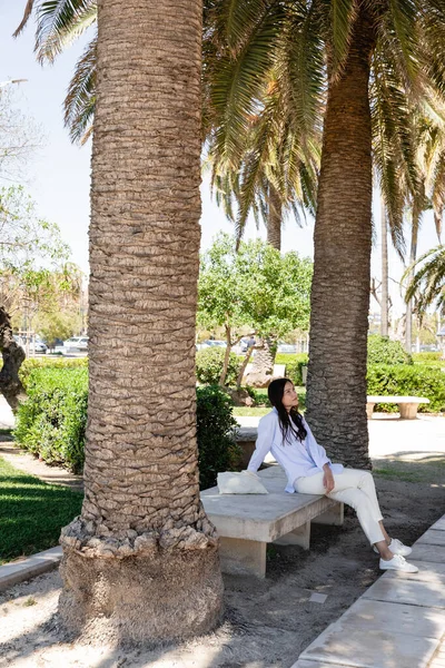 Vue pleine longueur de la femme assise sur le banc dans le parc sous les paumes géantes — Photo de stock