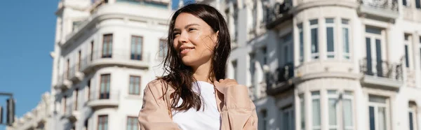 Positive woman looking away near white blurred buildings, banner — стоковое фото