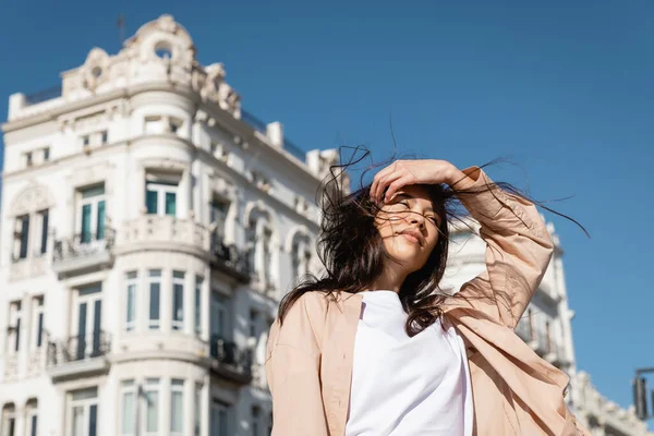 Low angle view of brunette woman fixing hair while standing on wind in city — Stock Photo