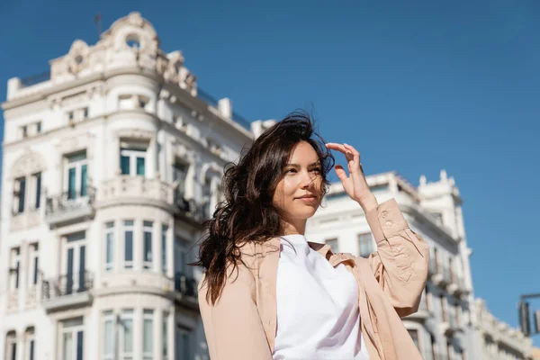 Brünette Frau steht auf windiger Straße in der Nähe weißer Gebäude auf verschwommenem Hintergrund — Stockfoto