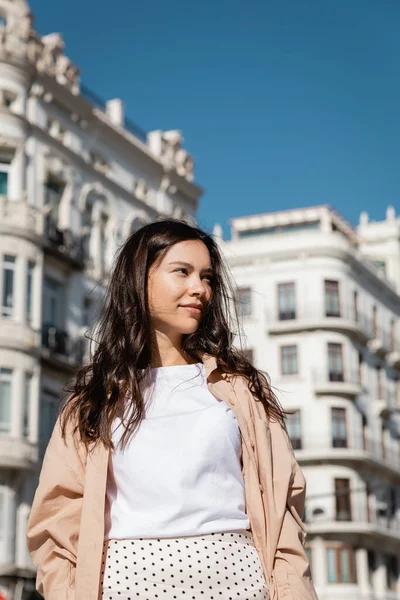 Brunette woman smiling while standing on urban street and looking away — стоковое фото