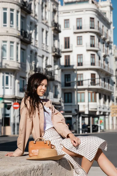 Stylish brunette woman sitting on street near straw hat and handbag — Stock Photo