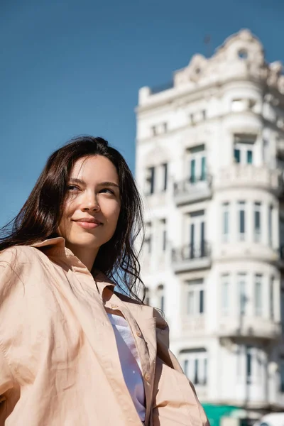 Brunette woman in beige shirt smiling near white building on blurred background — Foto stock